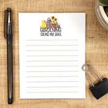 Load image into Gallery viewer, image of lined notepad on wooden background. words Gardening calms my soul with an image of boots, watering can, potted sunflower, and digger is above the words.
