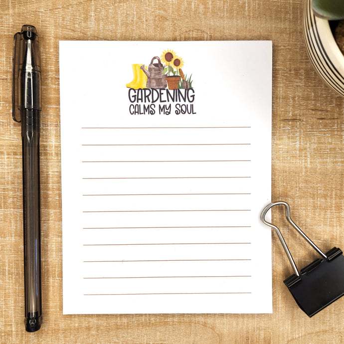 image of lined notepad on wooden background. words Gardening calms my soul with an image of boots, watering can, potted sunflower, and digger is above the words.